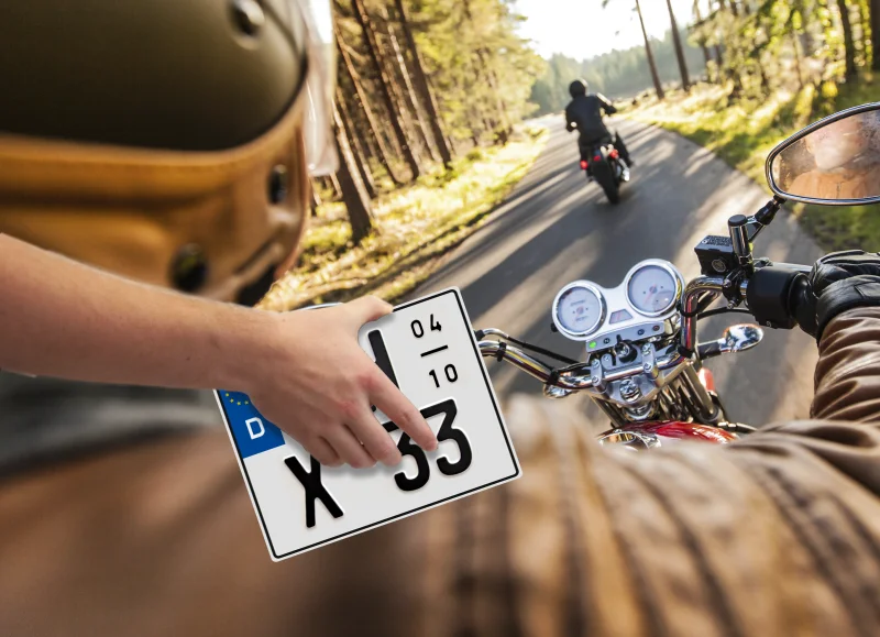 Zwei Motorräder befahren Straße durch Wald, man blickt hinterem Motorradfahrer über die Schulter, man sieht auf Lenker mit Tacho, eine Hand hält ein Motorrad Saisonkennzeichen ins Bild.