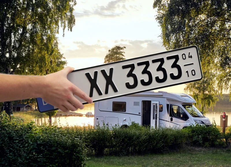 Zwischen Wiese, Büschen und 2 großen Bäumen steht ein weißes Wohnmobil mit geöffneter Tür. Im Vordergrund hält eine Hand ein Saisonkenneichen mit der Zeichenfolge XX 3333 04-10.